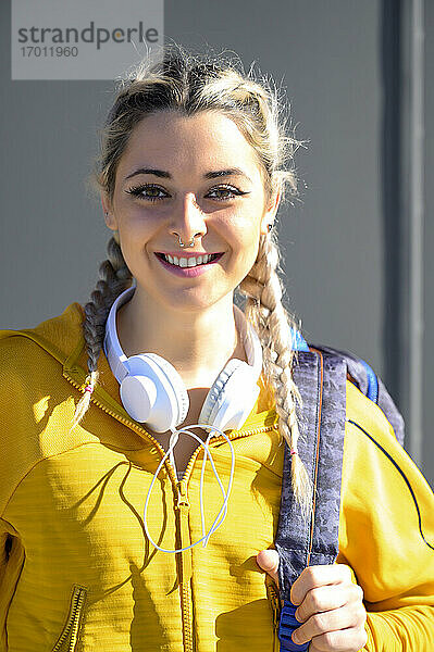 Lächelnde Sportlerin mit Rucksack und Kopfhörern steht im Freien