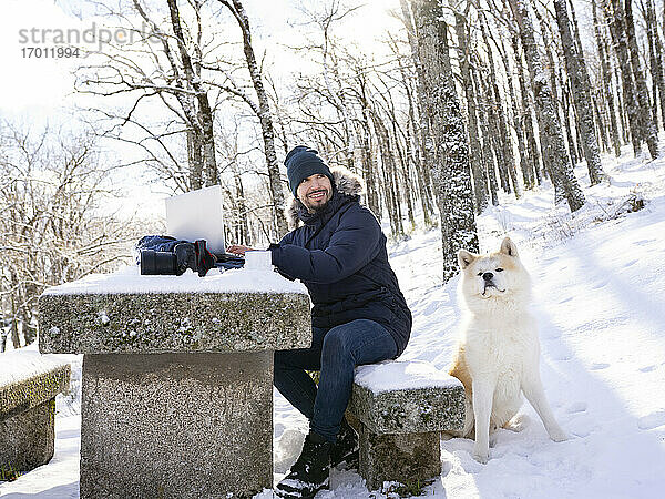 Fröhlicher Fotograf mit Laptop  der wegschaut  während er auf einer Bank neben einem Akita-Hund im Schnee sitzt