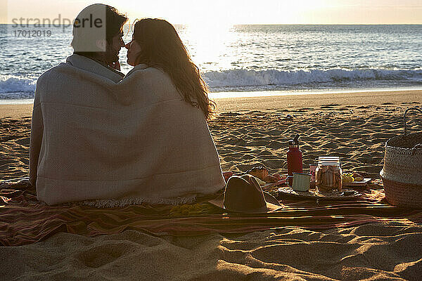 Paar in Decke gehüllt  das sich am Strand sitzend die Nase reibt