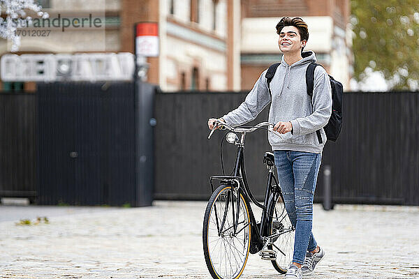 Lächelnder  modischer junger Mann mit Rucksack  der auf einem Fußweg in der Stadt Fahrrad fährt