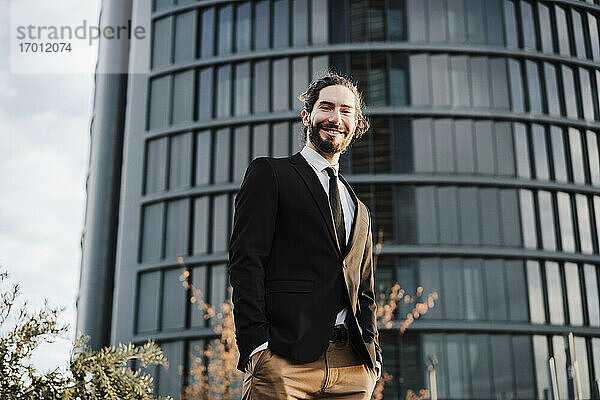 Junger Geschäftsmann mit Händen in den Taschen  der lächelnd vor einem Gebäude steht