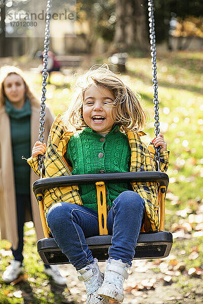 Lächelndes Mädchen  das auf einer Schaukel spielt  mit einer im Hintergrund stehenden Frau im Park