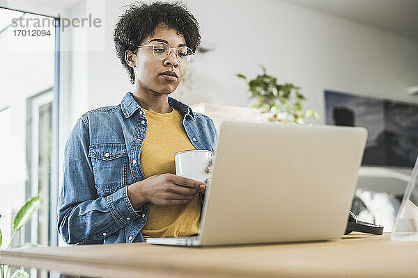 Frau hält Kaffeetasse und schaut auf Laptop zu Hause