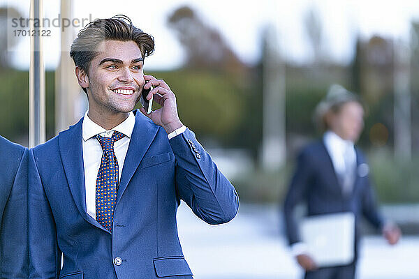Glücklicher Geschäftsmann im Anzug  der sich an ein Glas lehnt  während er mit seinem Smartphone telefoniert