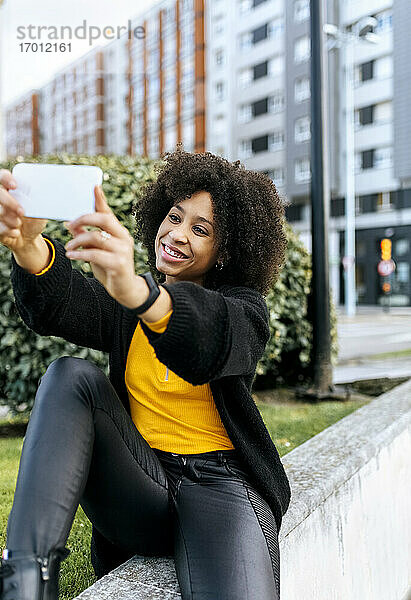 Lächelnde schöne Frau  die ein Selfie mit dem Mobiltelefon macht  während sie auf einem Geländer in der Stadt sitzt