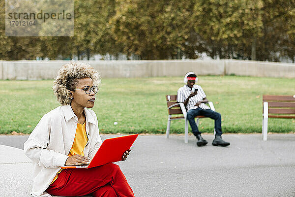 Frau  die einen Laptop benutzt  während sie auf einer Bank sitzt  mit einem Freund im Hintergrund auf der Bank