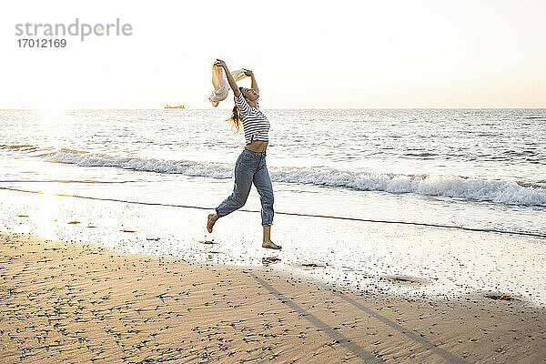 Unbeschwerte junge Frau mit Jacke beim Laufen am Strand gegen den klaren Himmel bei Sonnenuntergang
