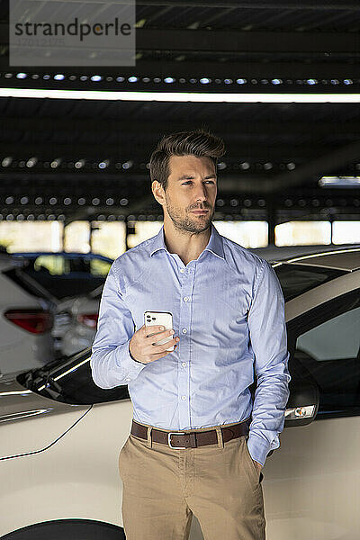 Männlicher Unternehmer mit Hand in der Tasche  der ein Mobiltelefon hält  während er auf einem Parkplatz steht