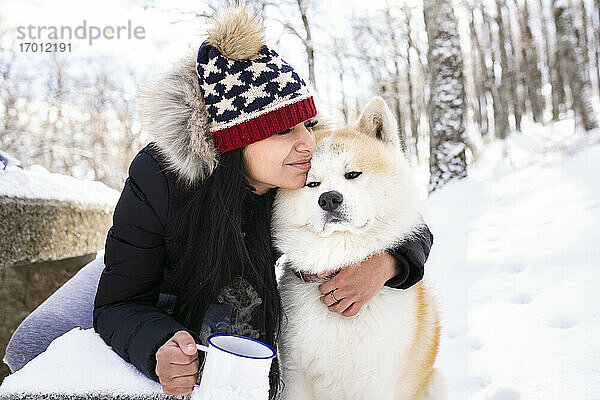 Frau mit geschlossenen Augen umarmt Akita-Hund und hält Kaffeetasse im Winter