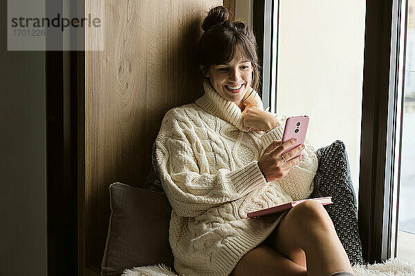 Frau im Pullover lächelt bei einem Videotelefonat mit einem Mobiltelefon am Fenster zu Hause