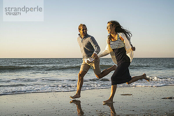 Fröhliches junges Paar  das sich beim Laufen am Strand während des Sonnenuntergangs an den Händen hält