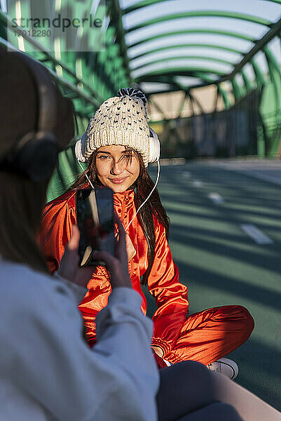 Teenager  der auf einer Brücke sitzend seinen Freund mit dem Handy fotografiert