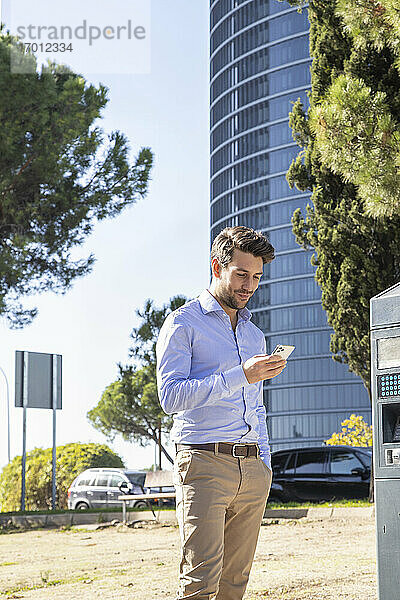Geschäftsmann  der ein Smartphone benutzt  während er an einer Parkuhr in der Stadt steht