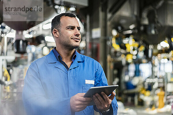 Ein lächelnder Techniker schaut weg  während er ein digitales Tablet in der Industrie hält
