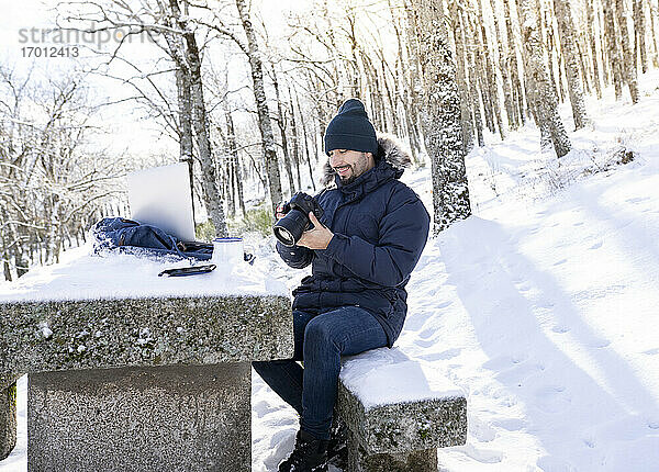 Lächelnder Fotograf mit Kamera auf einer Felsenbank im Wald sitzend bei Schneewetter