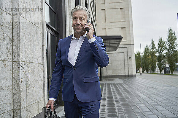 Älterer männlicher Unternehmer  der mit seinem Handy telefoniert  während er an einem Gebäude in der Stadt vorbeigeht