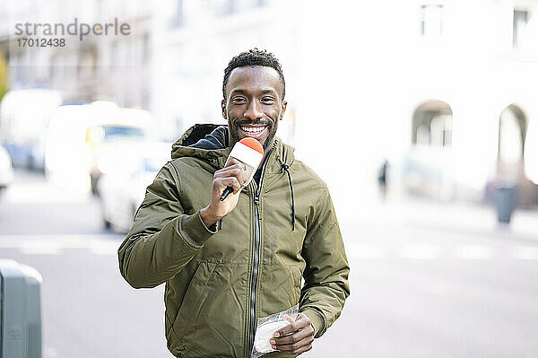 Lächelnder männlicher Journalist mit Mikrofon auf der Straße in der Stadt