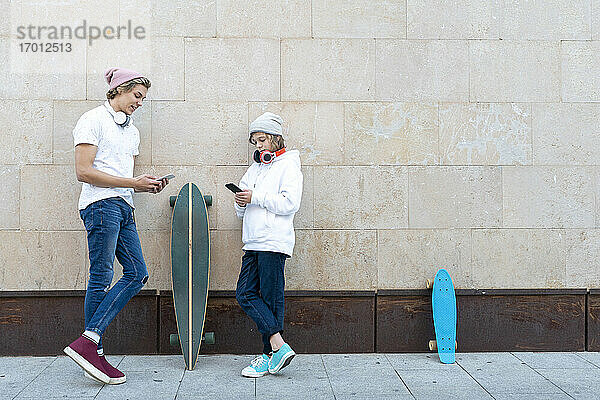Junger Mann und Junge mit Strickmütze  der ein Mobiltelefon benutzt  während er mit dem Skateboard an der Wand steht