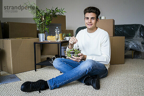 Lächelnder junger Mann mit Salat im Wohnzimmer einer neuen Loftwohnung
