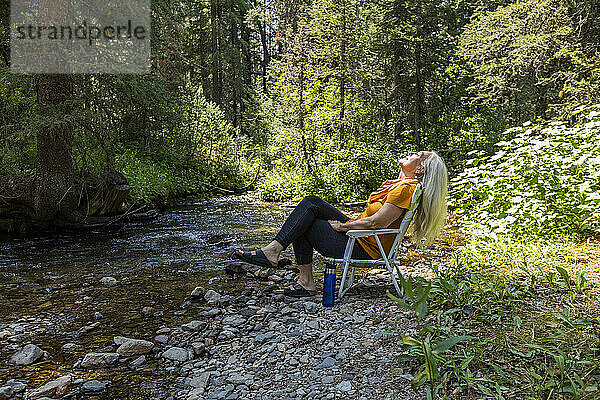 USA  Idaho  Sun Valley  Frau sitzt auf Stuhl am Flussufer im Wald