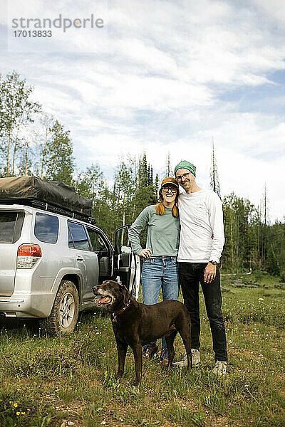 USA  Utah  Uinta National Park  Portrait eines lächelnden Paares mit Hund auf einer Wiese stehend  Geländewagen im Hintergrund