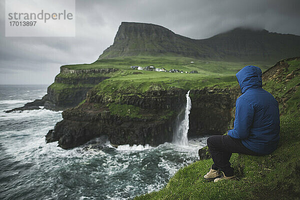 Dänemark  Färöer Inseln  Gasadalur Dorf  M-Lafossur Wasserfall  Mann sitzt am Rande der Klippe und schaut auf Mulafossur Wasserfall
