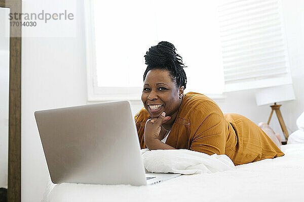 Porträt einer lächelnden Frau auf dem Bett liegend vor einem Laptop