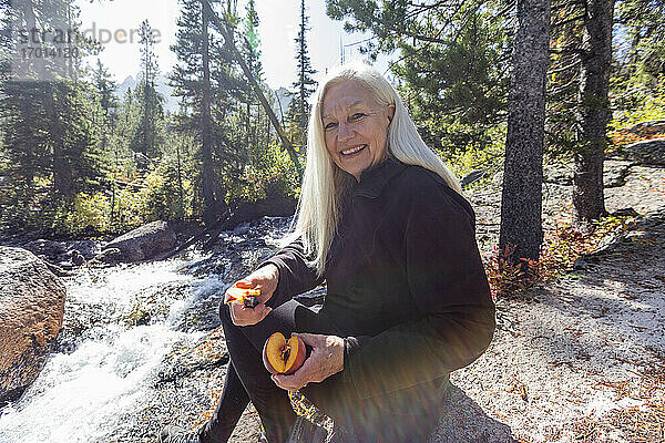 USA  Idaho  Stanley  Senior weiblichen Wanderer lächelnd und Schneiden frischen Pfirsich neben dem Bach