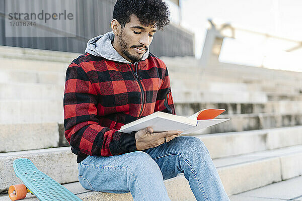 Junger Mann liest ein Buch  während er auf einer Treppe neben einem Skateboard sitzt