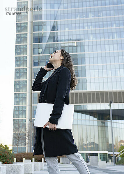 Geschäftsfrau mit Laptop  die lächelt  während sie mit einem Mobiltelefon spricht  beim Spaziergang in der Stadt