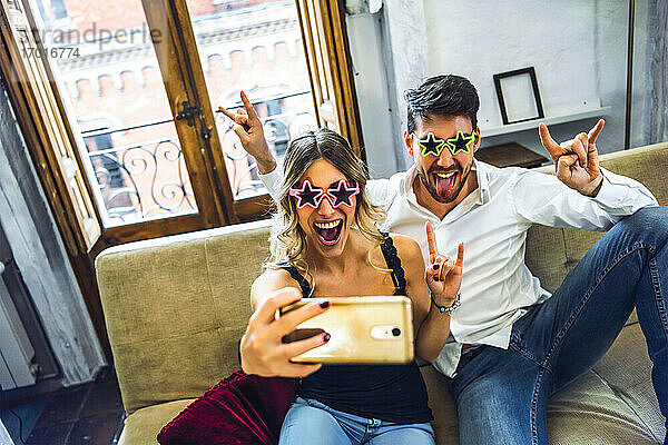 Verspieltes junges Paar  das eine Comedy-Brille trägt und ein Selfie auf der Couch im heimischen Wohnzimmer macht