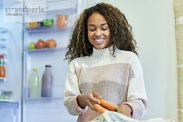 Lächelnde junge Frau  die in der Küche zu Hause Karotten aus einem Netzbeutel nimmt