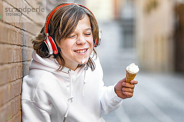 Lächelnder Junge mit Kopfhörern  der ein Eis in der Hand hält und sich an die Wand lehnt