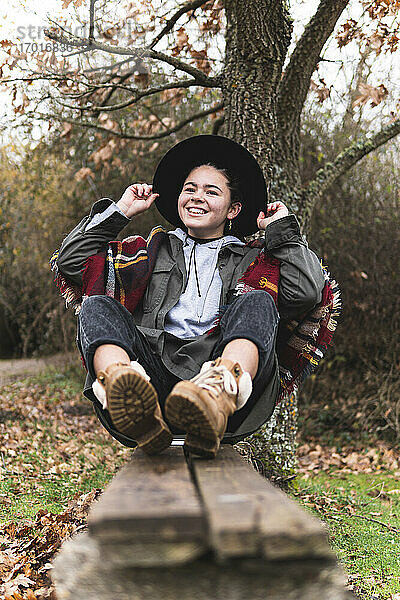 Lächelndes Teenager-Mädchen mit Hut  das auf einer Planke in einer Herbstlandschaft sitzt