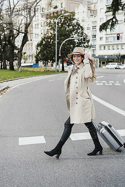 Glückliche Frau mit Gepäck auf Rädern  die einen Hut hält  während sie auf einer Straße in der Stadt spazieren geht