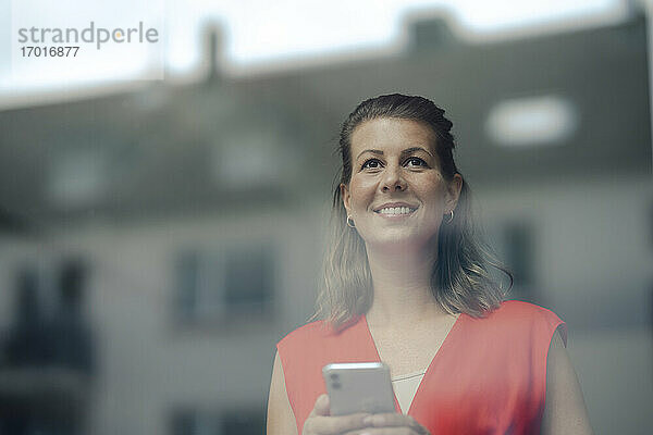 Lächelnde Geschäftsfrau mit Smartphone schaut durch ein Fenster