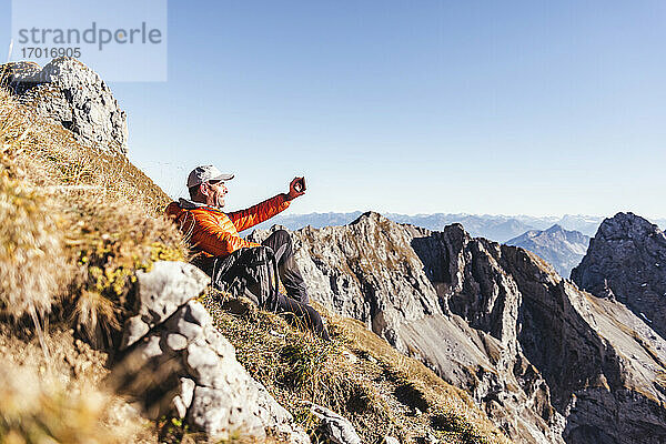 Älterer Mann  der ein Selfie mit seinem Smartphone macht  während er auf einem Berg gegen den klaren Himmel sitzt