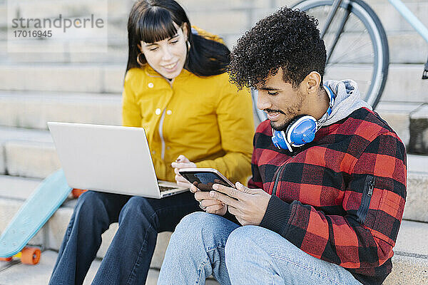 Junger Mann  der ein digitales Tablet benutzt  während er neben einer Freundin sitzt  die einen Laptop auf einer Treppe benutzt
