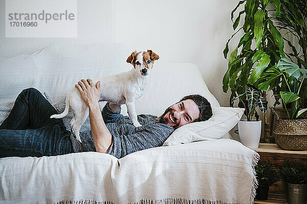 Glücklicher Mann lächelt  während er mit seinem Haustier spielt  das auf dem Sofa zu Hause liegt