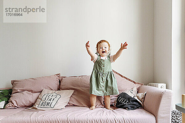 Lächelndes kleines Mädchen  das mit erhobenen Armen auf dem Sofa steht