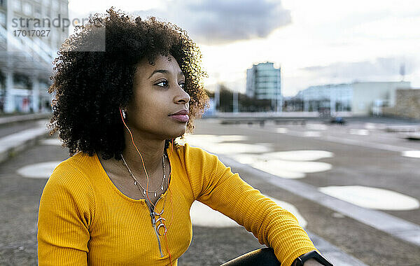 Nachdenkliche Frau mit Afro-Haar hört Musik  während sie gegen den Himmel in der Stadt sitzt