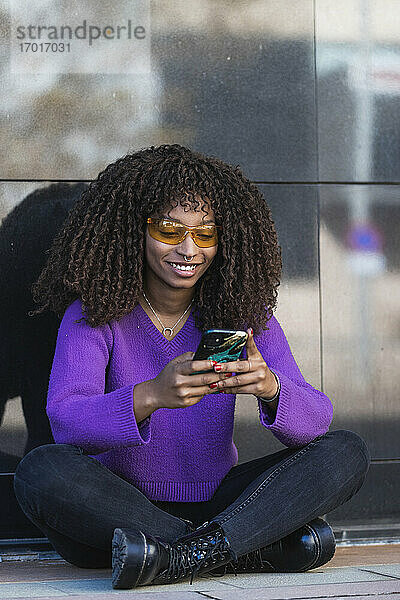 Lächelnde Frau auf dem Bürgersteig sitzend  während sie ein Mobiltelefon an der Wand benutzt