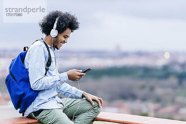 Lächelnder junger Mann hört Musik und benutzt sein Smartphone auf einer Terrasse gegen den Himmel