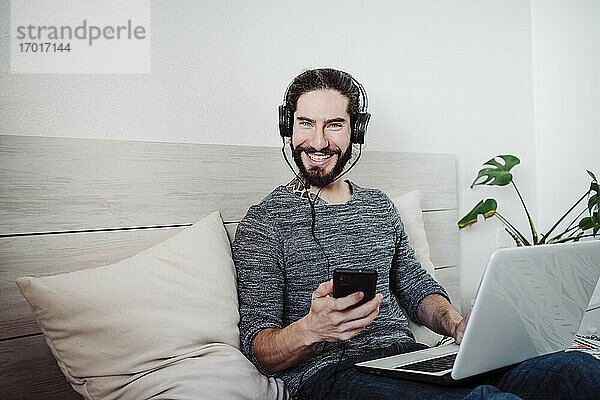 Lächelnder Mann mit Kopfhörern  der ein Mobiltelefon benutzt  während er mit einem Laptop auf dem Bett zu Hause sitzt