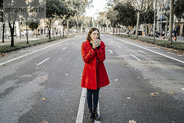 Modische Frau in roter Jacke hält Kaffeetasse mit geschlossenen Augen auf der Straße
