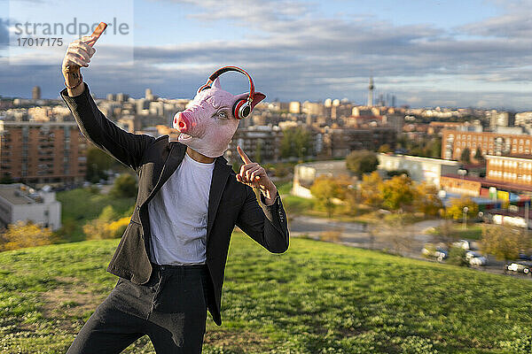 Geschäftsmann trägt Schweinemaske und drahtlose Kopfhörer  während er ein Selfie mit seinem Smartphone auf einem Hügel macht