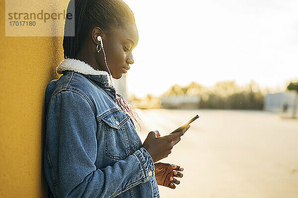 Teenager-Mädchen mit Smartphone beim Hören von Musik durch gelbe Wand auf der Straße