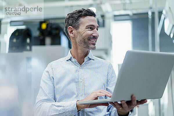 Lächelnder männlicher Fachmann mit Laptop  der in einer Fabrik wegschaut