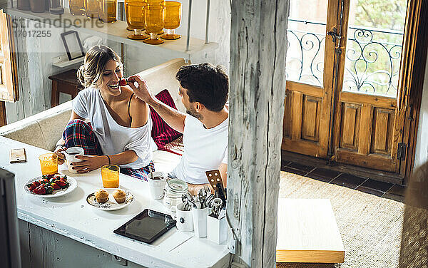 Junger Mann füttert Freundin mit Erdbeeren beim Frühstück zu Hause