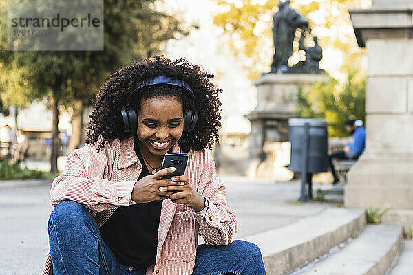 Glückliche junge Frau mit Afro  die ein Mobiltelefon benutzt  während sie auf einer Treppe in der Stadt Musik hört